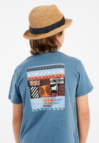 Camiseta corta de manga azul con un vibrante diseño gráfico en la parte posterior que incluye olas, patrones y texto. Un sombrero de paja añade textura y contraste de color.