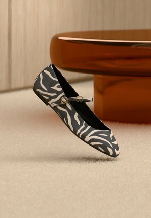 Fekete-fehér zebra csíkos lapos cipő arany csattos pánttal, szőnyeggel borított padlón, egy barna lekerekített asztalláb mellett pihenve.