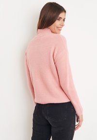 Maglione rosa chiaro realizzato in morbido tessuto testurizzato, con colletto alto e orlo a coste, abbinato a jeans neri. Vista da dietro.