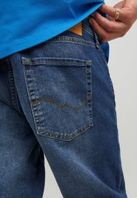 Vaqueros de mezclilla en azul oscuro con una vista en primer plano del bolsillo trasero, que presenta costuras amarillas y una etiqueta de cuero marrón claro cerca de la cinturilla.