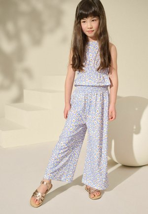 Jumpsuit floral bleu clair avec un haut à volants, une taille élastique, des jambes larges et un motif de fleurs jaunes et blanches. Associé à des sandales dorées.