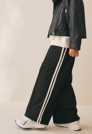 Person iført sorte bukser med vide ben og hvide striber, hvide sneakers, en hvid sweatshirt og en cropped sort læderjakke.