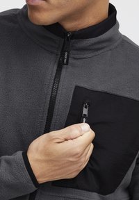 Chaqueta de forro polar gris con un bolsillo con cremallera negra. Presenta un alto cuello y una cremallera frontal. Tejido texturizado, hardware de cremallera resistente.