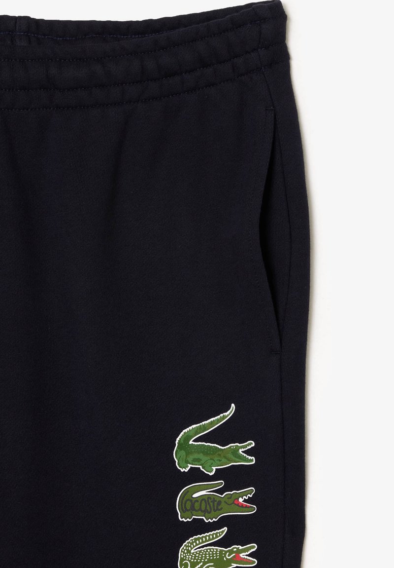 Pantalons de survêtement noirs avec une taille élastique et trois logos de crocodile verts empilés verticalement près de la poche droite.