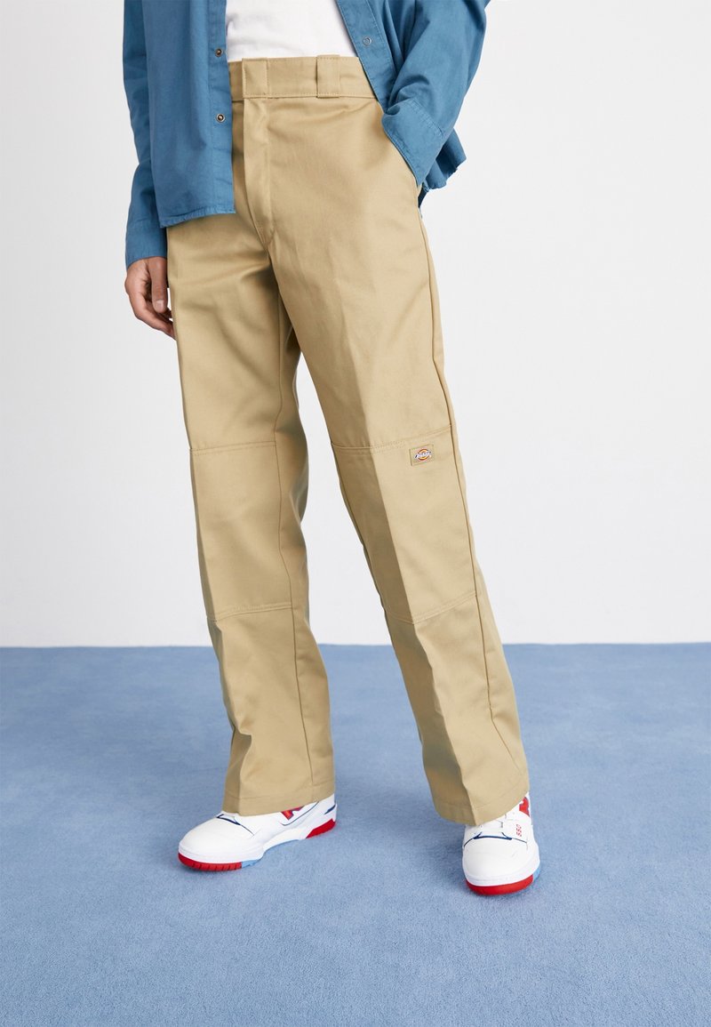 Pantalon de travail beige à coupe droite, avec plis sur le devant, genoux renforcés et un petit patch logo. Associé à des baskets blanches avec des accents rouges.