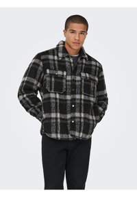 Jeune homme portant une veste en polaire à carreaux noirs et gris avec deux poches poitrine et un pantalon noir, les mains dans les poches.