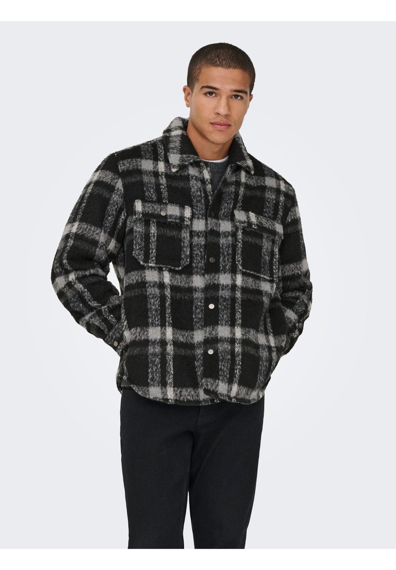 Jeune homme portant une veste en polaire à carreaux noirs et gris avec deux poches poitrine et un pantalon noir, les mains dans les poches.