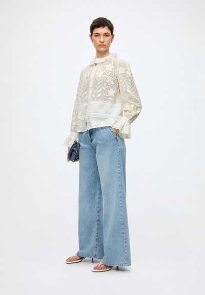 Femme portant un chemisier en dentelle florale crème transparente, un jean large bleu clair, des sandales blanches à talons, tenant un petit sac bleu marine.
