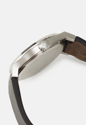 Silberner Edelstahl-Uhr mit rundem Gehäuse, ausgestattet mit einer Drückerkronen und einem schwarzen Lederarmband mit glatter Textur.