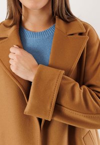 Cappotto color cammello con una trama morbida, caratterizzato da ampi rever e dettagli cuciti, indossato sopra un maglione a coste di un delicato blu.