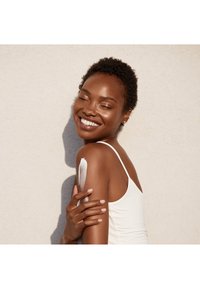 En person med kort, mörkt hår bär en vit linne och applicerar lotion på sin arm. Hyn är slät och glödande mot en ljus bakgrund.