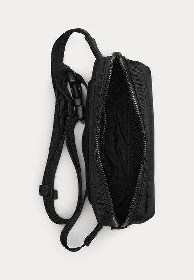 Melnas auduma jostas soma ar regulējamu siksnu un atvērtu galveno rāvējslēdzēja nodalījumu, kurā redzama auduma oderējuma iekšpuse.