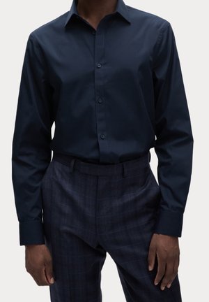 Homme portant une chemise bleu foncé à manches longues boutonnée rentrée dans un pantalon habillé bleu foncé à carreaux avec une ceinture.