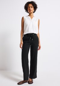 Bílý bezrukávový top s výstřihem do V a dvěma kapsami na prsou, kombinovaný s volnými černými kalhotami s elastickým pasem a šňůrkou.