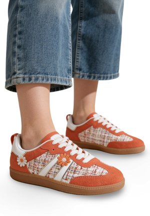 Pieds portant des baskets à motif floral orange et blanc avec des lacets blancs et des semelles en gomme, assorties à un pantalon en jean bleu raccourci.