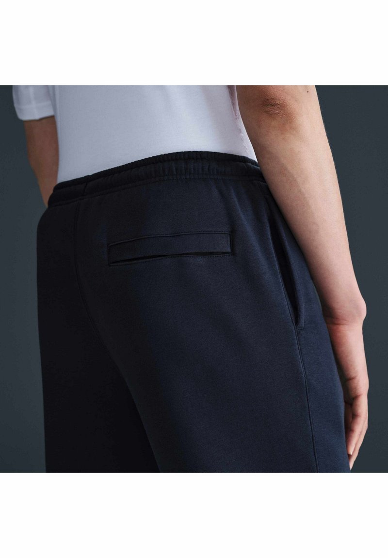 Persona che indossa joggers blu navy con cintura elastica e tasca posteriore, abbinati a una camicia bianca, vista di lato.