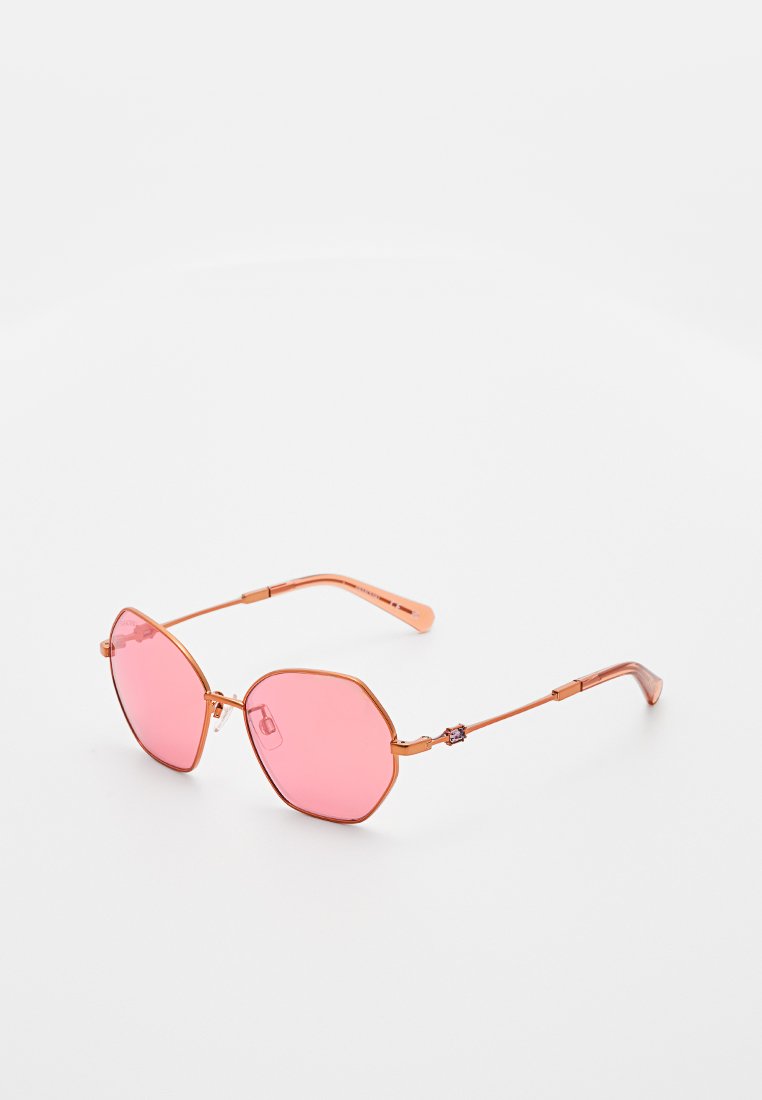 Zonnebril met dunne roségouden metalen monturen en roze geometrische lenzen, geplaatst op een witte ondergrond.