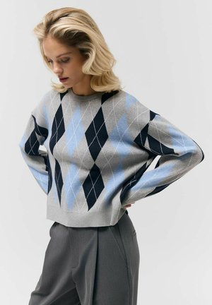 Kobieta o blond włosach, ubrana w szary, niebieski i czarny sweter w kratę oraz szare spodnie, stoi z rękami na biodrach na jednolitym tle.