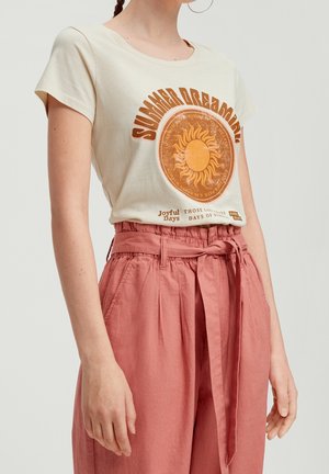 Vrouw draagt een crèmekleurig T-shirt met een oranje zon en de tekst "Summer Dreamin'", gecombineerd met een hoge taille, vastgebonden dusty roze broek.