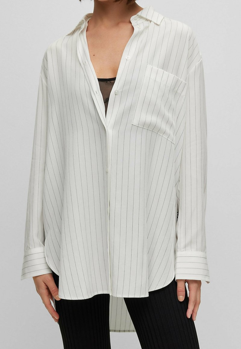 Femme portant une chemise blanche oversize à fines rayures noires verticales, col ouvert, poche poitrine, et pantalon noir côtelé.