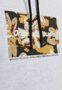 Sudadera con capucha de color gris claro que presenta un patrón rectangular floral en amarillo, negro y marrón, con el texto "Originals Studio" y cordones negros.