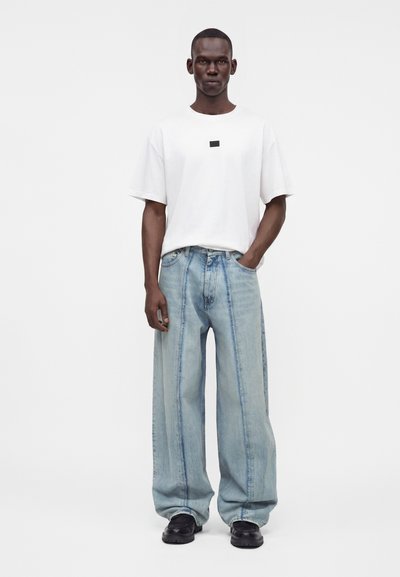 Άνδρας που φοράει ένα απλό λευκό υπερμεγέθη T-shirt και φαρδιά ανοιχτό μπλε τζιν, στέκεται μπροστά σε ένα λευκό φόντο.
