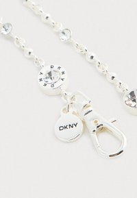 Sudraba tonis pērļu ķēde ar caurspīdīgiem kristāliem, ar DKNY apaļo piekariņu, mazu DKNY birku un omārdzīgo aizdari uz balta fona.