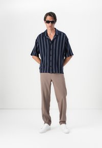 Camisa de manga corta a rayas en azul marino con botones, pantalones beige y zapatillas blancas. Los accesorios incluyen gafas de sol negras. Fondo minimalista.