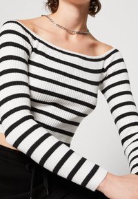 Haut à rayures horizontales noir et blanc, côtelé et décolleté épaule dénudée. Les manches longues et la coupe ajustée mettent en avant un look contemporain.