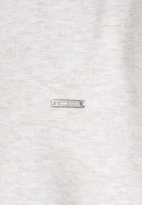 Petite étiquette métallique rectangulaire avec "SKILODOX" gravé, fixée sur un tissu beige clair.