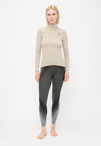 Lys beige langermet topp med kvart glidelås og logo, sammen med svarte leggings som har en gradienttekstur fra solid til flekket.