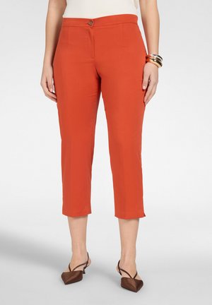 Donna che indossa pantaloni arancioni corti a gamba dritta, décolleté marroni con tacco a gattino e punta appuntita, top bianco e braccialetti impilati su un polso.