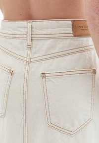 Valkoinen denim-hame, jossa on oranssit ompeleet, takataskut niiteillä ja vaaleanruskea nahkaleima. Siisti muotoilu, keskikorkea istuvuus.