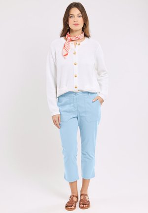 Femme portant un cardigan blanc boutonné, un pantalon court bleu clair, des sandales marron et un foulard à rayures rouges autour du cou, debout avec une main dans la poche.