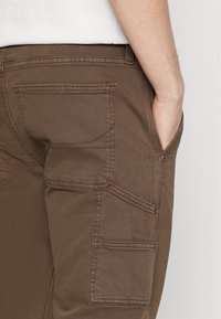 Ruskeat kestävät cargo-housut, joissa on useita taskuja ja hienovaraiset ompelut. Sivutaskussa vetoketjulisä.