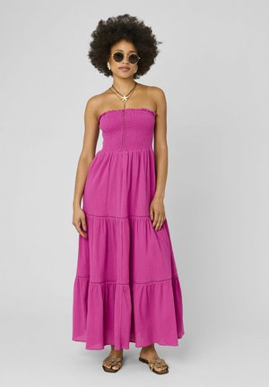 Femme avec afro portant des lunettes de soleil rondes, une robe longue rose à volants sans bretelles, des colliers superposés et des sandales fleuries, debout devant un fond uni.