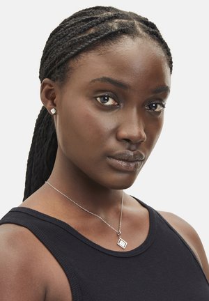 Silberne Halskette mit einem geometrischen Anhänger, getragen zu einem schwarzen ärmellosen Oberteil. Das Model hat geflochtenes Haar und dez normales Makeup.