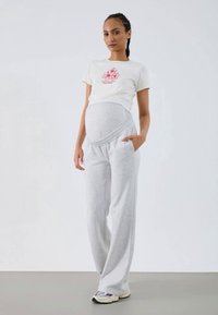 Umstandsmode bestehend aus einer hellgrauen, dehnbaren Sweatpants mit Taschen und einem beigefarbenen T-Shirt, das mit einem pinken floralen Design verziert ist.