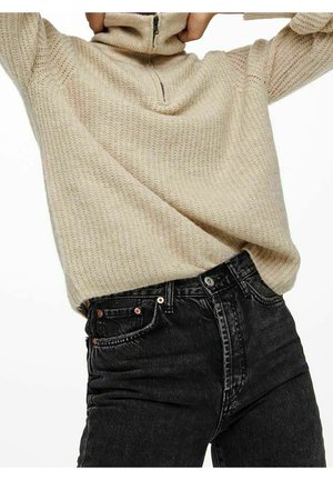 Beige striktrøje med halv lynlås krave, parret med mørke sorte jeans med knapdetaljer og en tætsiddende pasform.
