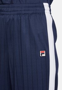 Pantaloni tuta a righe blu scuro con pannelli laterali bianchi e un piccolo logo ricamato. Realizzati in tessuto testurizzato; presentano una vita elastica.