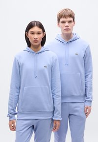 Ανοιχτό μπλε φούτερ με κουκούλα από απαλό ύφασμα, με τσέπες καγκουρό και ελαστικές μανσέτες. Μικρό πράσινο λογότυπο στο αριστερό στήθος.