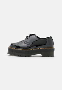 Scarpa di vernice nera con punta tonda, suola in gomma testurizzata spessa, cuciture gialle e design con lacci.