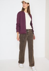 Giacca viola con zip e orlo regolabile, maglione crema e pantaloni larghi marroni. Sneakers bianche con accenti rossi; stile casual.