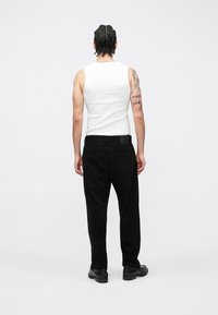 Λευκό αμάνικο τοπ και μαύρο τζιν με ίσια γραμμή, εμφανιζόμενο από πίσω. Φοράει μαύρα παπούτσια, έχει πλεξούδες στα μαλλιά και εμφανίζονται τατουάζ.