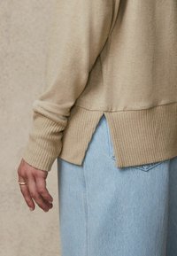 Beige neulepaita, jossa on ribbireunat ja halkiohelma, yhdistettynä vaaleansinihin farkkuihin. Lähikuvassa näkyy tekstuuri.
