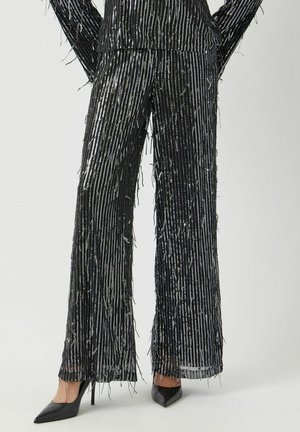 Pantaloni neri a gamba larga con strisce verticali metalliche, caratterizzati da dettagli in frange lungo i bordi. Abbinati a tacchi a punta neri.
