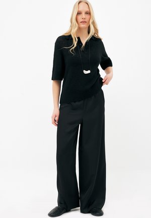 Blonde vrouw die een zwart kort mouwtruitje, zwarte wijde broek, zwarte instapschoenen en een witte hangerketting draagt, staand met hand op de heup.