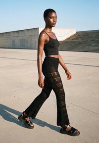 Femeie îmbrăcată cu top negru fără mâneci și pantaloni largi cu model, mergând în aer liber pe beton, aproape de scări, sub un cer senin.