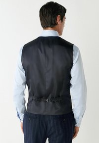 Zwarte satijnen vest met een gladde textuur en een verstelbaar riemdetail aan de achterkant. Draag het over een lichtblauw shirt met lange mouwen en een gestreepte broek.