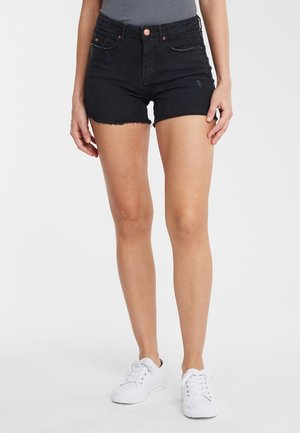 Kvinder ben iført sorte denimshorts med frynsete kanter, hvide sneakers og en grå top mod en ensfarvet lys baggrund.